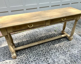 May include: A light brown wooden console table with two drawers and brass-colored handles. The table has four turned legs and a lower stretcher. The table is placed on a patterned rug.