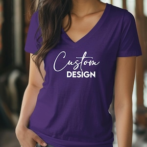 May include: A purple V-neck t-shirt with the words "Custom DESIGN" in white script. The shirt is made of a soft material and has short sleeves. The shirt is a solid color and is suitable for casual wear.