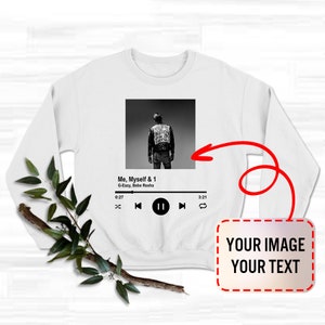 May include: White sweatshirt with a black and white image of a person wearing a leather jacket. The image is a music player screen with the song title "Me, Myself & 1" by G-Eazy and Bebe Rexha. The music player has a play button, a pause button, and a forward button. The sweatshirt has a blank space for a custom image and text.