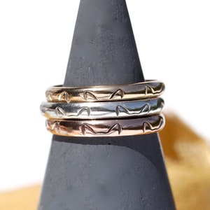 May include: Three stacking rings in different metal finishes: rose gold, silver, and gold. Each ring has a repeating pattern of small, stylized cat faces.