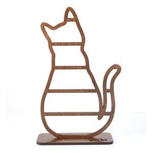 May include: A wooden cat-shaped earring holder in a warm brown hue. The holder features multiple horizontal bars with small holes for displaying earrings. The base is rectangular, and the design includes a tail and a cutout head.