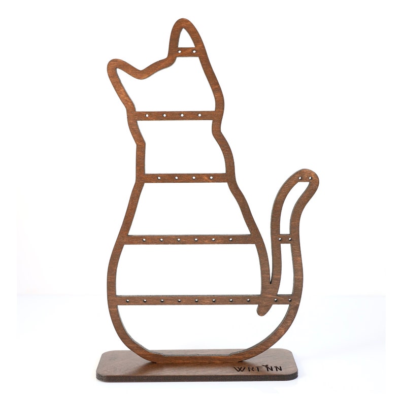 May include: A wooden cat-shaped earring holder in a warm brown hue. The holder features multiple horizontal bars with small holes for displaying earrings. The base is rectangular, and the design includes a tail and a cutout head.