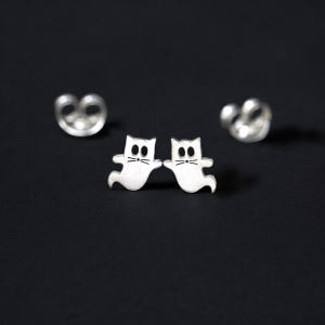 May include: A pair of silver cat ghost stud earrings. The earrings are small and feature a simple design of a cat with its eyes and mouth open, as if it is a ghost.