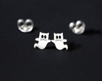 Pode incluir: Um par de brincos de pressão de prata em forma de fantasma de gato. Os brincos são pequenos e apresentam um design simples de um gato com os olhos e a boca abertos, como se fosse um fantasma.