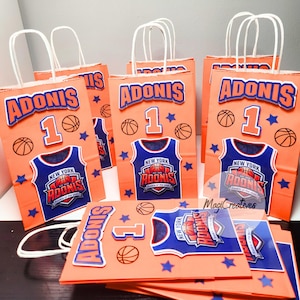 May include: Orange party favor bags with white handles, decorated with basketball-themed graphics. Each bag features the name "ADONIS", the number "1", a basketball jersey design, basketballs, and blue stars. The bags are designed for a birthday party.