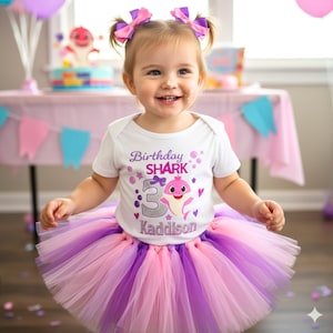May include: A young child wearing a white onesie with "Birthday Shark" text and a pink shark graphic, paired with a layered pink, purple, and lavender tutu. The child has pink and purple bows in their hair.
