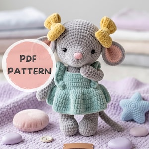 Op de afbeelding: Een gehaakte grijze muispop met een lichtblauwe jurk en gele strikken. De pop heeft roze oren en een roze neus. De afbeelding bevat de tekst "PDF PATTERN" in een roze cirkel.
