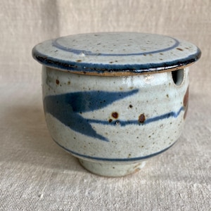 May include: A ceramic butter dish with a lid. The dish is off-white with blue and brown accents. The lid has a blue ring around the edge. The dish has a cylindrical shape with a small foot.