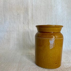 May include: A brown ceramic jar with a wide mouth and a smooth, glazed finish. The jar is approximately 10 cm tall and 7 cm wide.