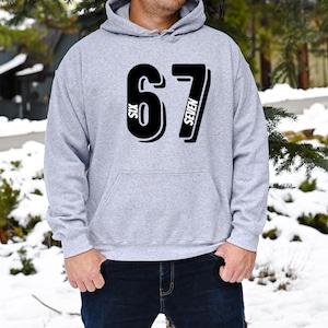 May include: A light gray hoodie with a large black graphic of the number "67" on the front. The word "SIX" is vertically printed on the left side of the number "6" and the word "SEVEN" is vertically printed on the right side of the number "7".