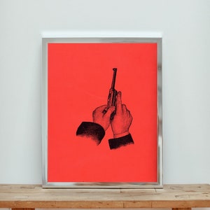 May include: A framed art print featuring a black and white illustration of hands holding a gun against a vibrant red background. The artwork is encased in a silver frame, creating a striking contrast. The print is displayed on a wooden surface.