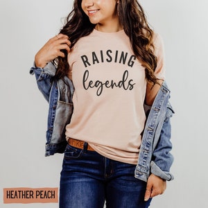 May include: Heather peach-colored t-shirt with the words "RAISING legends" in black script. The model is wearing a denim jacket and blue jeans. The shirt is a soft, casual style.