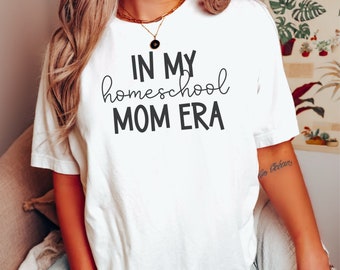 May include: White t-shirt with the text "IN MY homeschool MOM ERA" in black. The shirt is a casual style, likely made of cotton or a similar fabric. The text is in a mix of fonts, with the word "homeschool" in a cursive style.