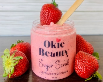 May include: A pink sugar scrub jar with a black label that reads "Okie Beauty Sugar Scrub Strawberrylicious 8.5 oz". The jar is surrounded by fresh strawberries.