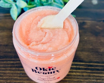 May include: A clear jar of pink sugar scrub with a wooden spoon inside. The jar has a label that says "Okie Beauty Luxury Skincare Sugar Scrub Pink Grapefruit 6.5 oz."