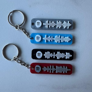 May include: Four keyrings in grey, blue, black, and red, each with a Spotify code design. The keyrings have a silver keyring and chain. The Spotify logo is white.