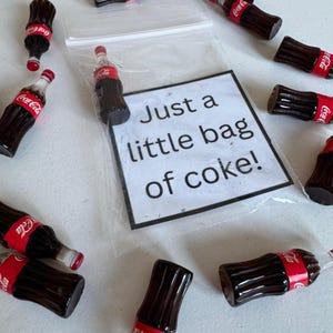 May include: A collection of miniature Coca-Cola bottles with red and white labels. The bottles surround a clear plastic bag with a sign saying "Just a little bag of coke!" The bottles are brown with red caps.