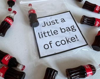May include: Miniature Coca-Cola bottles with red and white labels are scattered around a clear plastic bag. The bag contains a sign that reads "Just a little bag of coke!" The bottles are brown and have red caps.