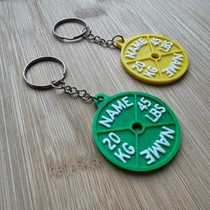 May include: Two weightlifting plate keyrings. One is yellow with white text, including "NAME", "20 KG", and "45 LBS". The other is green with white text, including "NAME", "20 KG", and "45 LBS". Both have silver keyrings.