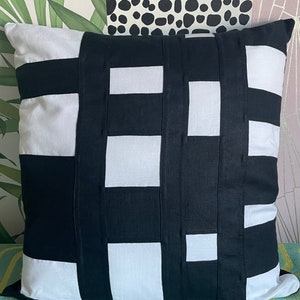 Puede incluir: Almohada de tiro con estampado geométrico en blanco y negro. La almohada está hecha de una tela suave y texturizada y tiene una forma cuadrada.