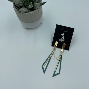 Può includere: Un paio di orecchini geometrici verdi e dorati con un design a triangolo. Gli orecchini sono su una carta nera con il testo "Mimi's World".