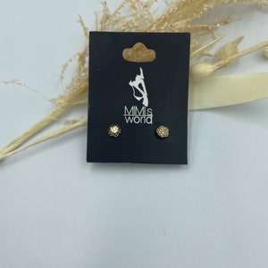 Puede incluir: Un par de pendientes de botón dorados con superficie texturizada. Los pendientes se muestran en una tarjeta negra con el texto "Mimi's World" impreso en ella.