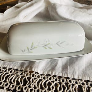 May include: A white porcelain butter dish with a lid, resting on a matching rectangular plate. The dish features a delicate floral design in pale green and gray. The plate has a gold trim.