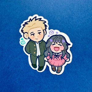 May include: A sticker featuring two cartoon characters, a male and a female, standing together. The male character wears a green jacket and the female character wears a pink dress. The sticker has a white border and is set against a blue background.