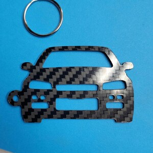 Puede incluir: Un llavero de metal negro y gris con forma de la parte delantera de un coche. El llavero tiene un patrón de fibra de carbono.