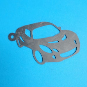 Pode incluir: Um berloque cinza escuro em forma de carro com um orifício circular na parte superior para pendurar. O berloque tem detalhes recortados para janelas e rodas, em um fundo azul brilhante.
