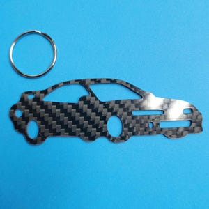 Könnte beinhalten: Ein schwarzer und silberner Autoschlüsselanhänger mit einem Karbonfaser-Muster. Der Schlüsselanhänger ist in Form eines Autos gestaltet und hat einen silbernen Ring, der daran befestigt ist.