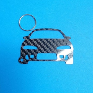 Może przedstawiać: Srebrny metalowy brelok do kluczy w kształcie samochodu z wzorem z włókna węglowego. Brelok ma srebrny pierścień przymocowany do góry.