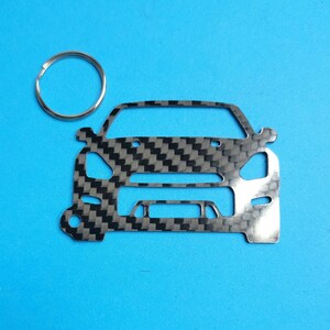 Puede incluir: Un llavero de metal plateado con un patrón de fibra de carbono. El llavero tiene la forma de la parte delantera de un coche y tiene un anillo plateado.