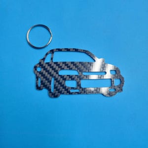 Puede incluir: Un llavero de coche plateado con un patrón de fibra de carbono. El llavero tiene la forma de un coche y tiene un anillo plateado adjunto.