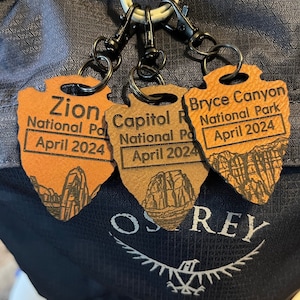 May include: Three brown leather keychains with a map design and the names of national parks: Zion National Park, Capitol Reef National Park, and Bryce Canyon National Park. Each keychain has the date "April 2024" engraved on it.