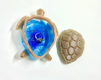 May include: A blue and brown resin turtle ring holder with a ring inside. The turtle is open and the top shell is off to the side.
