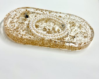 May include: Oval-shaped clear resin tray with gold leaf flakes embedded in the resin. A pearl necklace is resting in the tray.