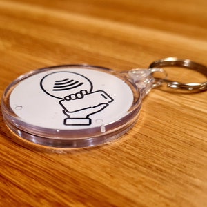 Pu&ograve; includere: Un portachiavi in plastica trasparente con un cerchio bianco che contiene un'icona in bianco e nero di una mano che tiene un telefono con un segnale wireless sopra.