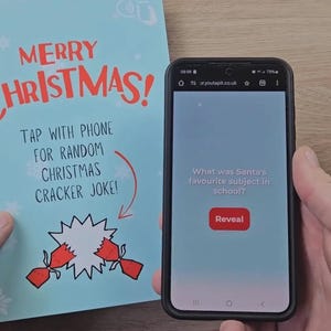 Może przedstawiać: Jasnoniebieska kartka z czerwonym napisem Merry Christmas i instrukcją dotknięcia telefonem w celu uzyskania świątecznego żartu, obok smartfona wyświetlającego stronę internetową z tekstem What was Santa's favourite subject in school?