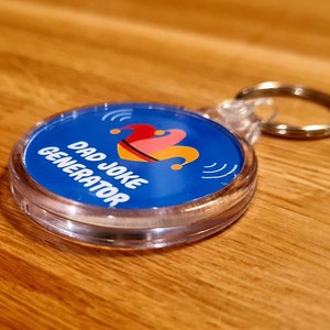 Pu&ograve; includere: Un portachiavi in plastica trasparente con un cerchio blu che contiene il testo "Dad Joke Generator" e un giullare dei cartoni animati con un cappello rosso, giallo e rosa. Il portachiavi ha un anello di metallo argentato.