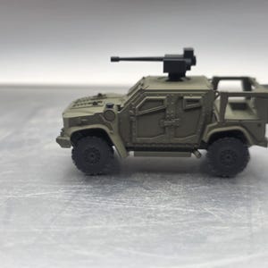 May include: A miniature olive drab military vehicle with a black gun turret on top. The toy truck has four black wheels and a detailed exterior, including windows and panels. The vehicle is designed to resemble a military transport.