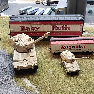 May include: Two toy military tanks, one larger than the other, are on a green grassy surface. The larger tank is tan and has a long barrel. The smaller tank is tan and has a shorter barrel. Behind the tanks are two toy train cars. The train car in the foreground is red and has the word "Bazooka" printed on it. The train car in the background is red and white and has the words "Baby Ruth" printed on it.