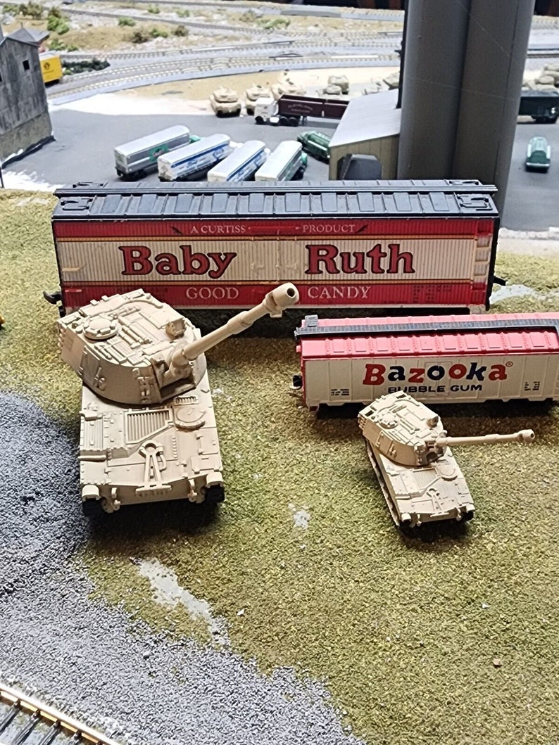 May include: Two toy military tanks, one larger than the other, are on a green grassy surface. The larger tank is tan and has a long barrel. The smaller tank is tan and has a shorter barrel. Behind the tanks are two toy train cars. The train car in the foreground is red and has the word "Bazooka" printed on it. The train car in the background is red and white and has the words "Baby Ruth" printed on it.