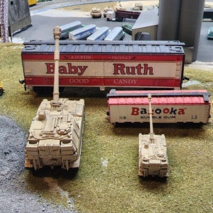 May include: A model train set with a red and white boxcar with the text "Baby Ruth Candy" on the side. There are two toy military tanks, one beige and one white, in the foreground.