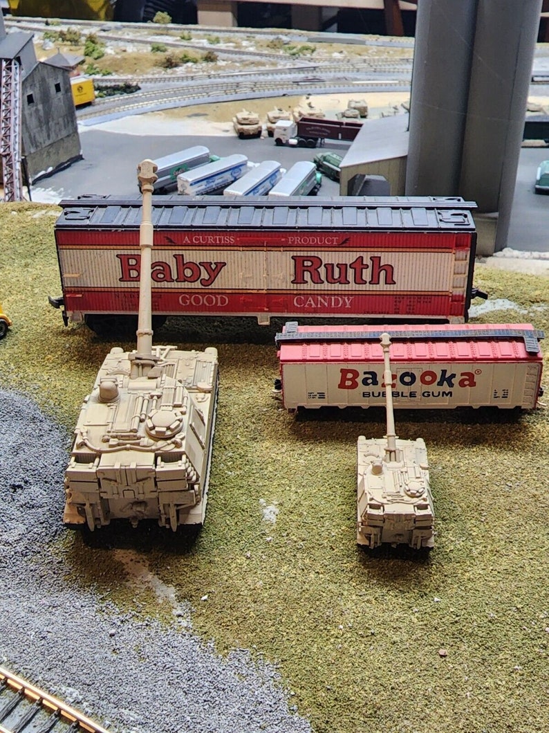 May include: A model train set with a red and white boxcar with the text "Baby Ruth Candy" on the side. There are two toy military tanks, one beige and one white, in the foreground.