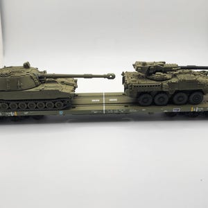 May include: A green military transport flatcar carrying two military vehicles. The vehicle on the left is a self-propelled howitzer, and the vehicle on the right is a light armoured vehicle.