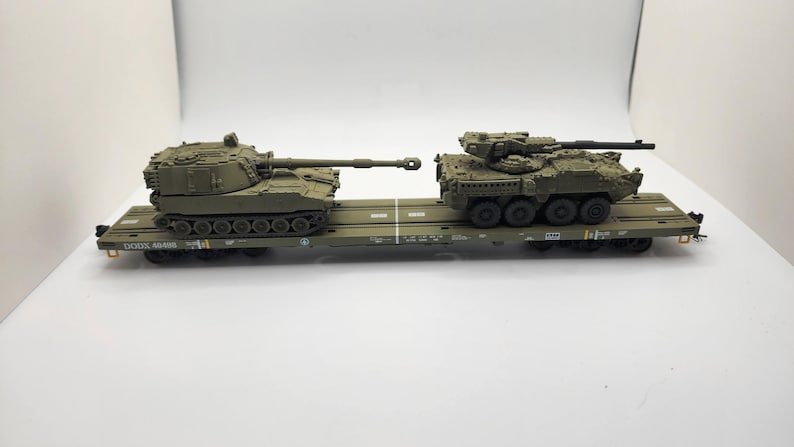 May include: A green military transport flatcar carrying two military vehicles. The vehicle on the left is a self-propelled howitzer, and the vehicle on the right is a light armoured vehicle.