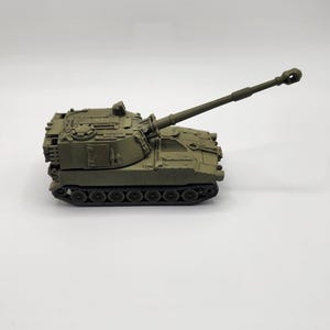 May include: A green toy model of a self-propelled howitzer artillery piece with a long barrel and a detailed design.