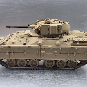 May include: A tan-colored scale model of an armored infantry fighting vehicle. The model features a rotating turret with a long black cannon, side-mounted storage boxes, a detailed hull with visible rivets, and a continuous black track system.