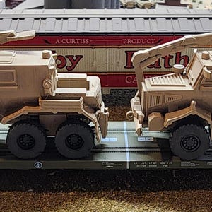 May include: Two tan military vehicles with cranes are being transported on a flatbed train car. The vehicles have a boxy design with a large cab and a flatbed in the back. The train car is labeled "DACOXX 4000".